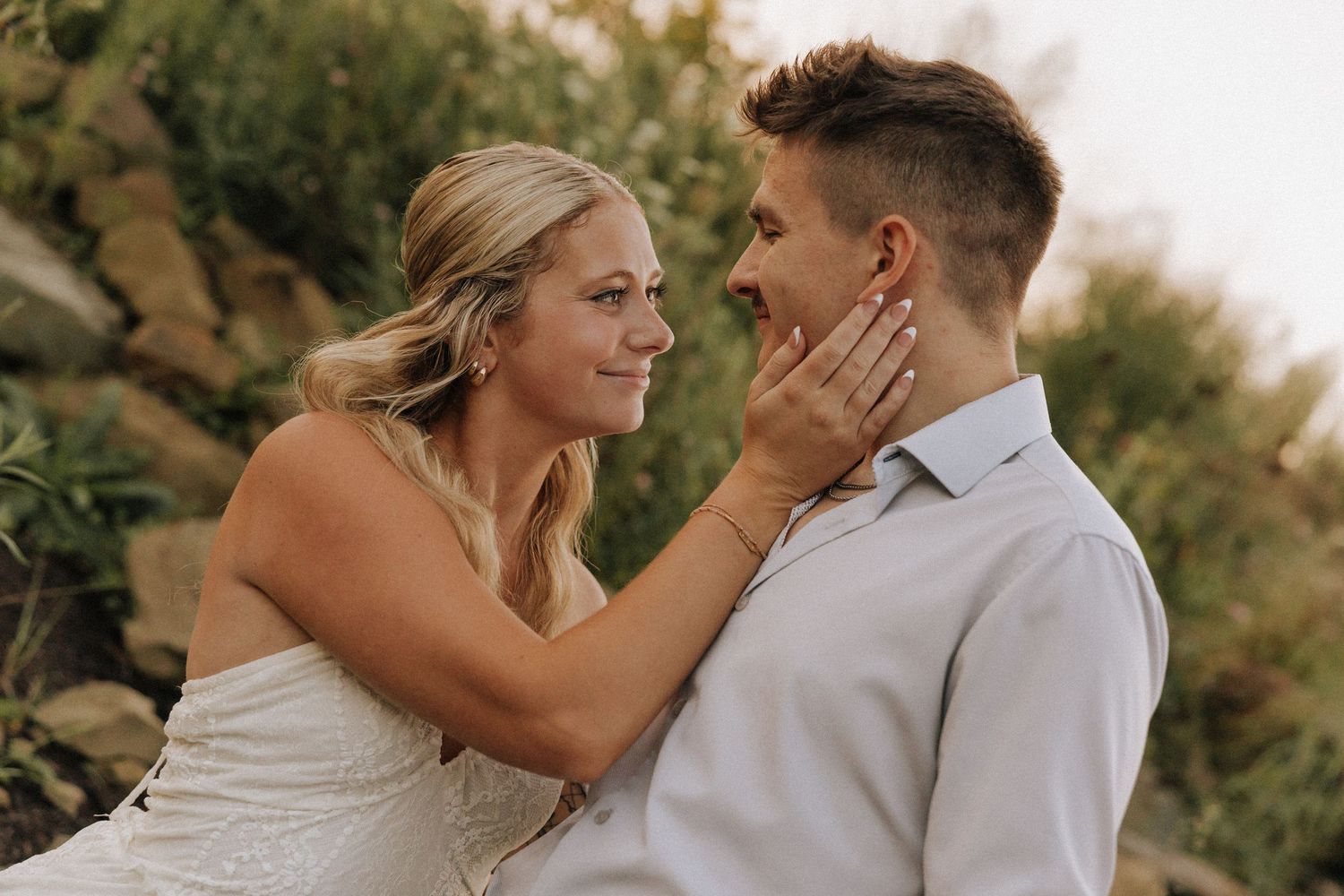 A bride tenderly holds her groom's face while smiling at him outdoors during a romantic wedding photo session.