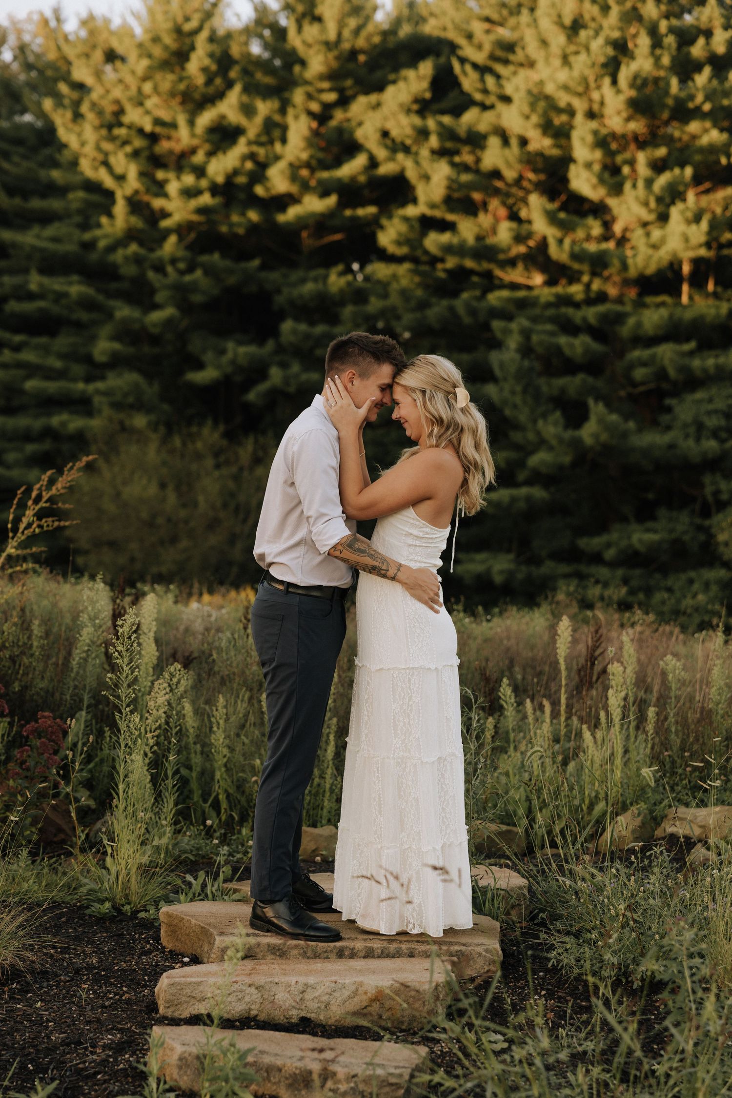 Couple in white dress and navy suit embrace on stone steps surrounded by lush greenery at golden hour.