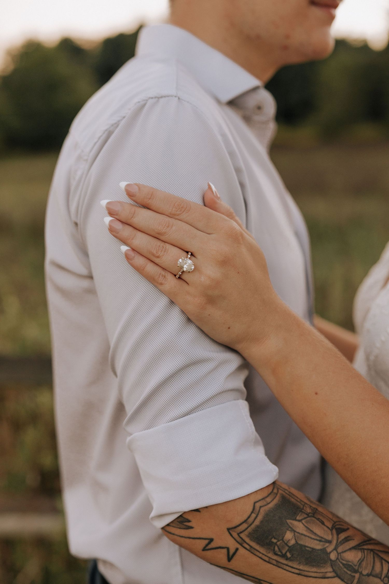Close-up of a woman's hand with an engagement ring resting on a man's shoulder, with a tattoo visible on her arm.