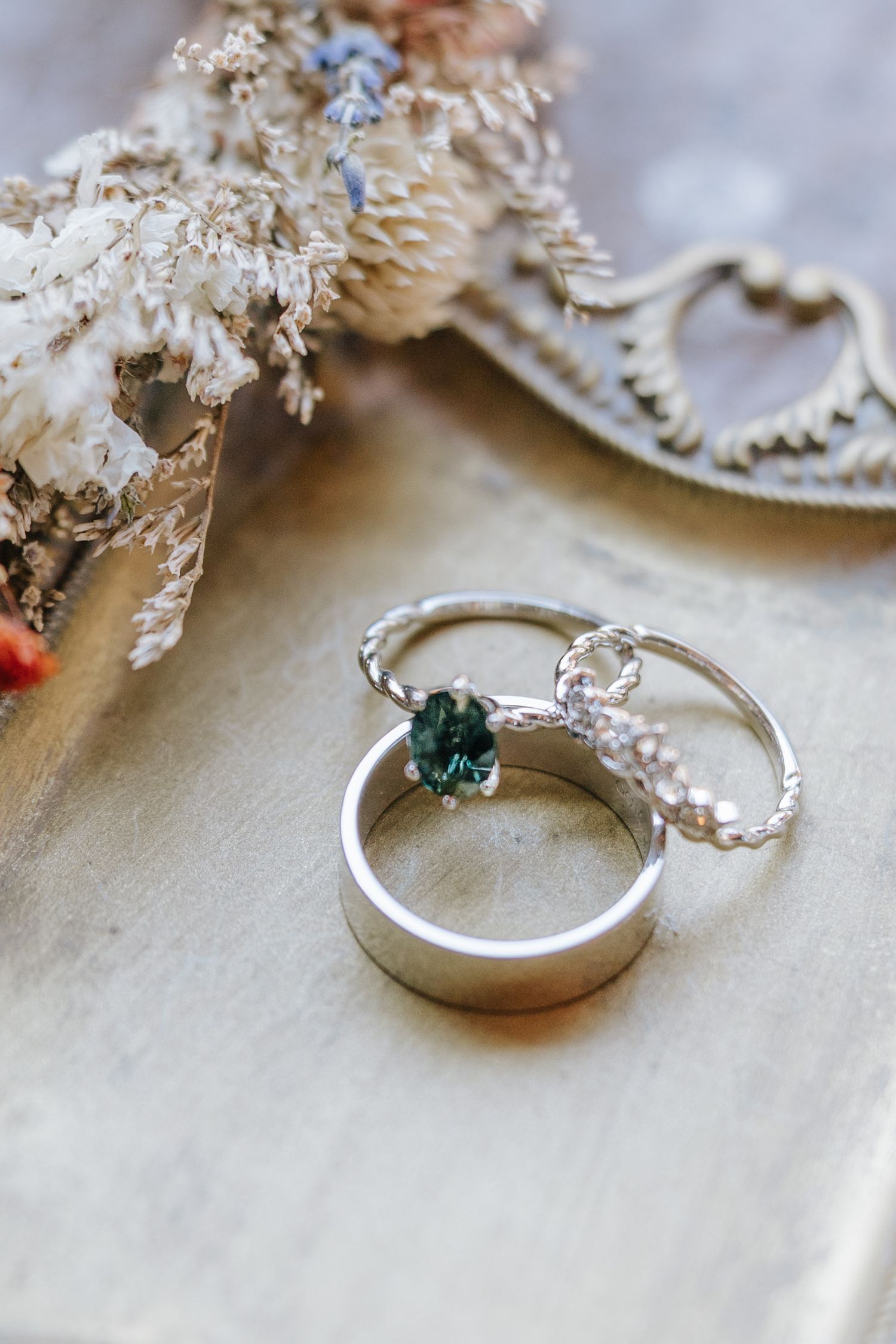 Delicate silver engagement ring with small green gemstone displayed on white surface with dried flowers.