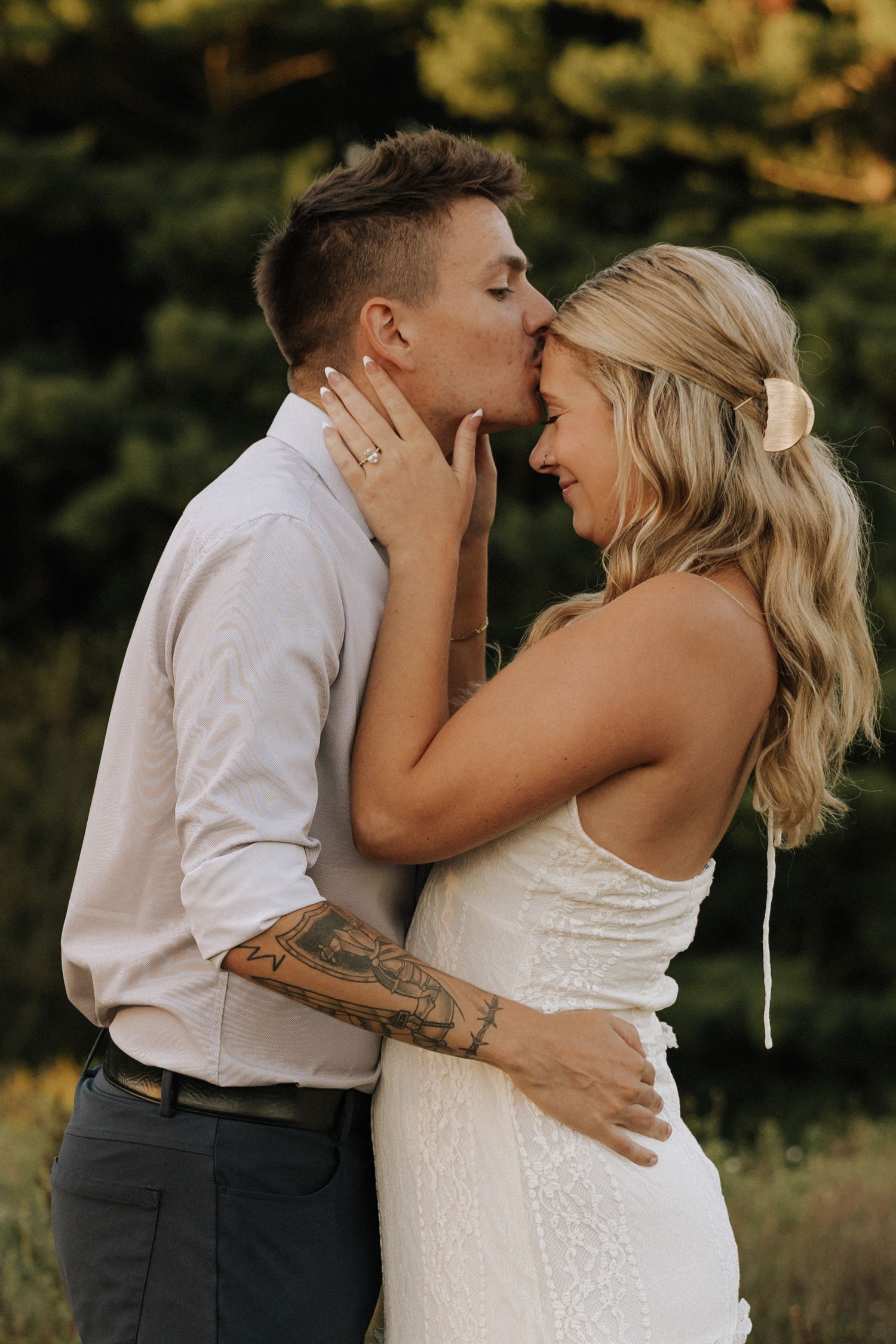 A couple shares a tender kiss outdoors, the groom in a white shirt embracing his blonde bride in a white dress.