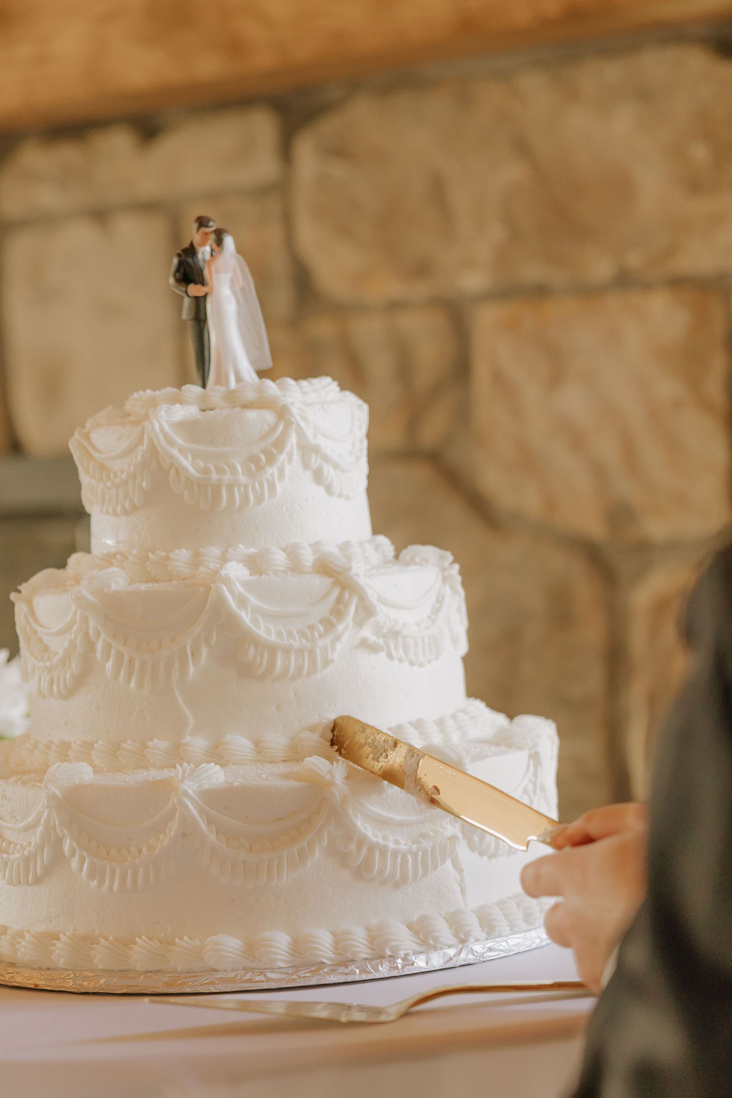 A white tiered wedding cake with delicate piped lace details and a traditional bride and groom topper being photographed.