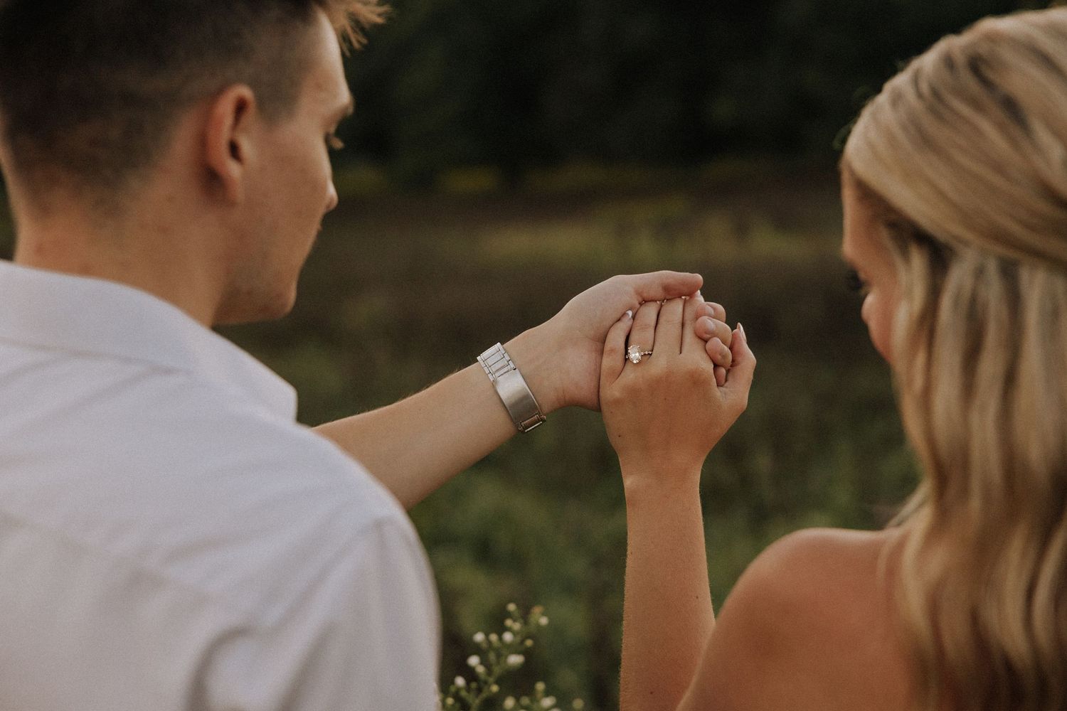 Couple holding hands outdoors, woman wearing engagement ring, romantic nature background.
