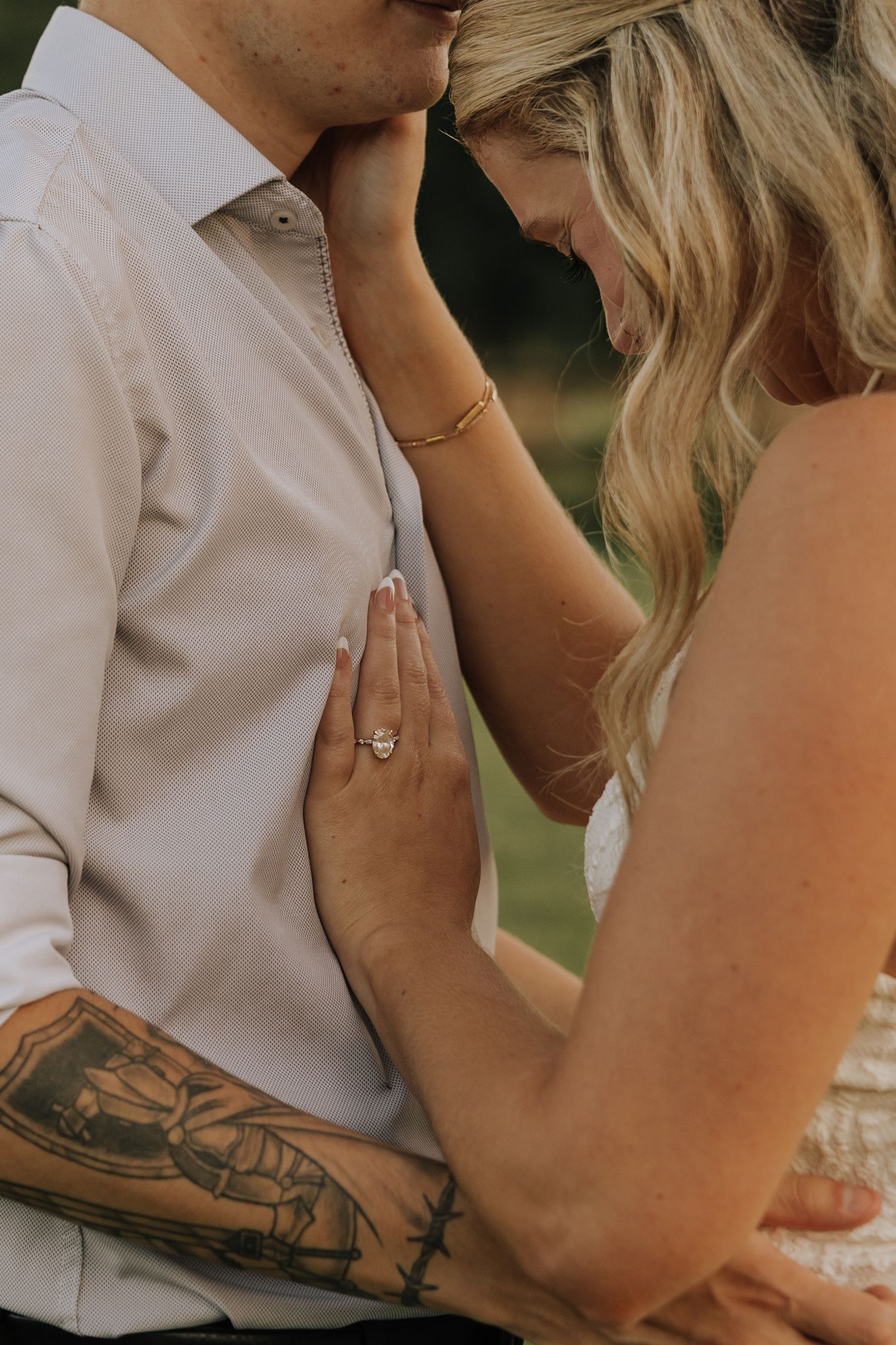 Couple sharing a romantic moment, woman with engagement ring and bracelet resting hand on mans chest, man with tattoo kissing her forehead.