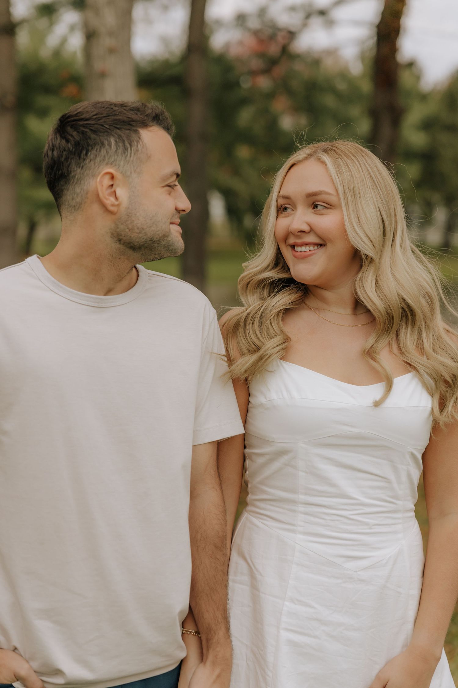A romantic outdoor portrait of a couple in white clothing smiling at each other.