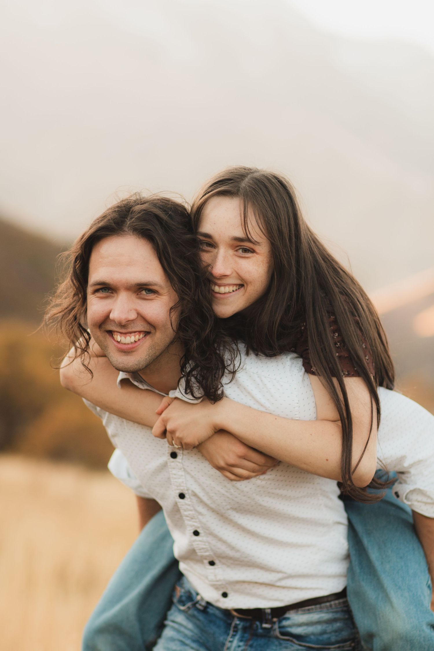 A joyful couple shares a piggyback ride moment outdoors against a warm autumn landscape.