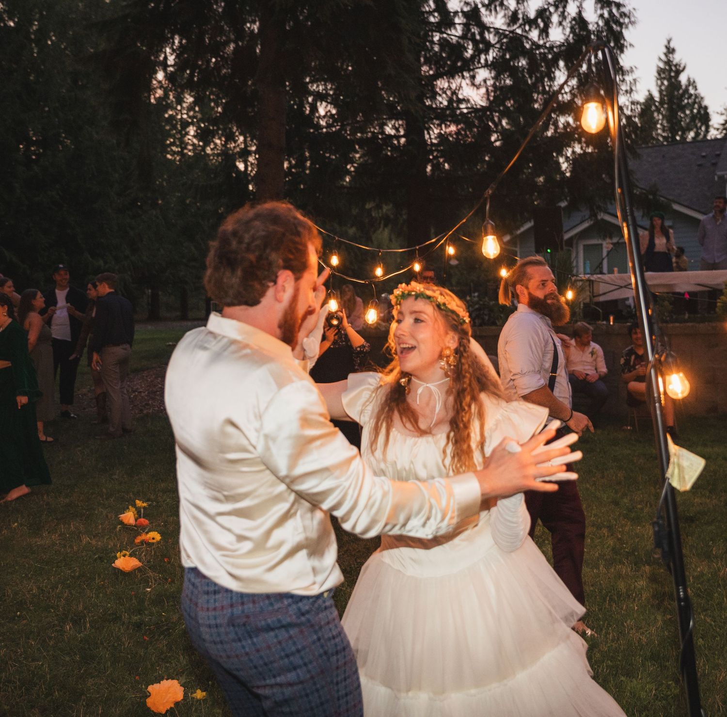 A couple dances together at dusk under string lights in a rustic outdoor wedding celebration.