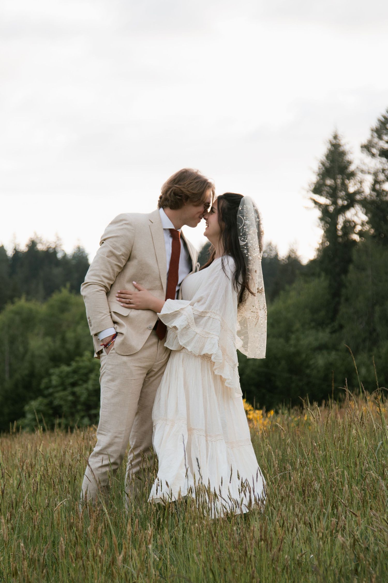 A romantic embrace captured in a lush meadow with evergreen trees in the background.