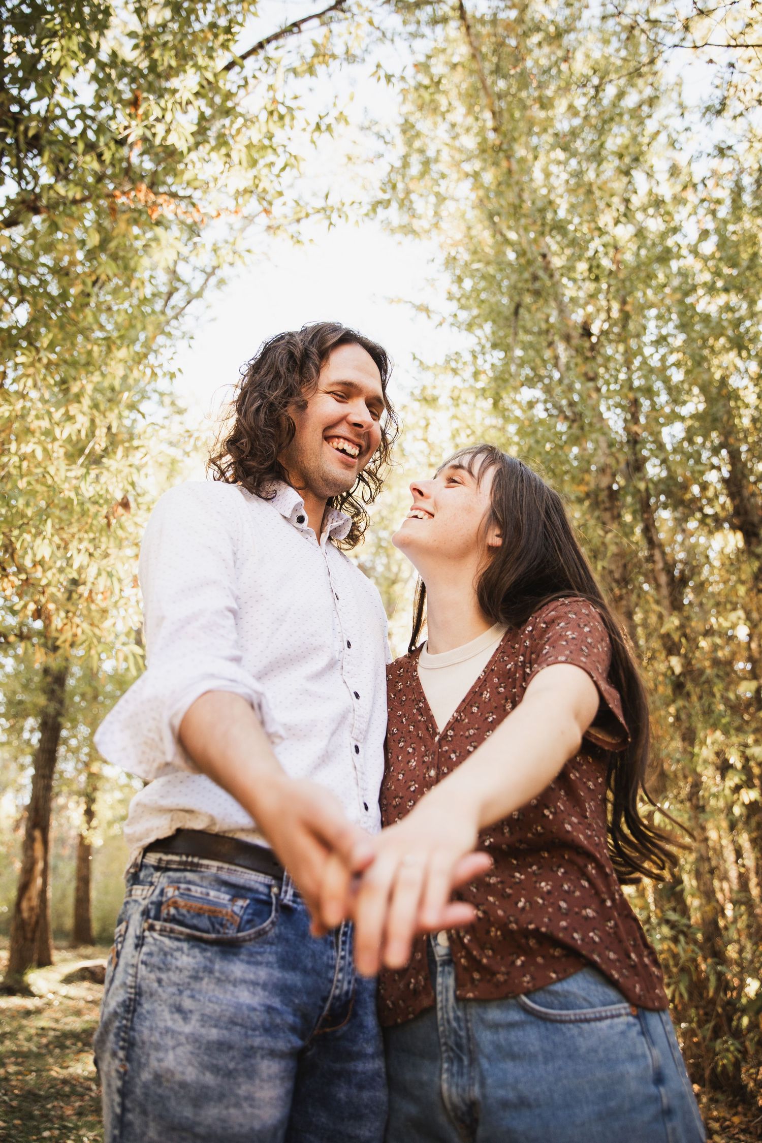 A couple laughs together while holding hands in a sunlit forest setting with golden light filtering through trees.