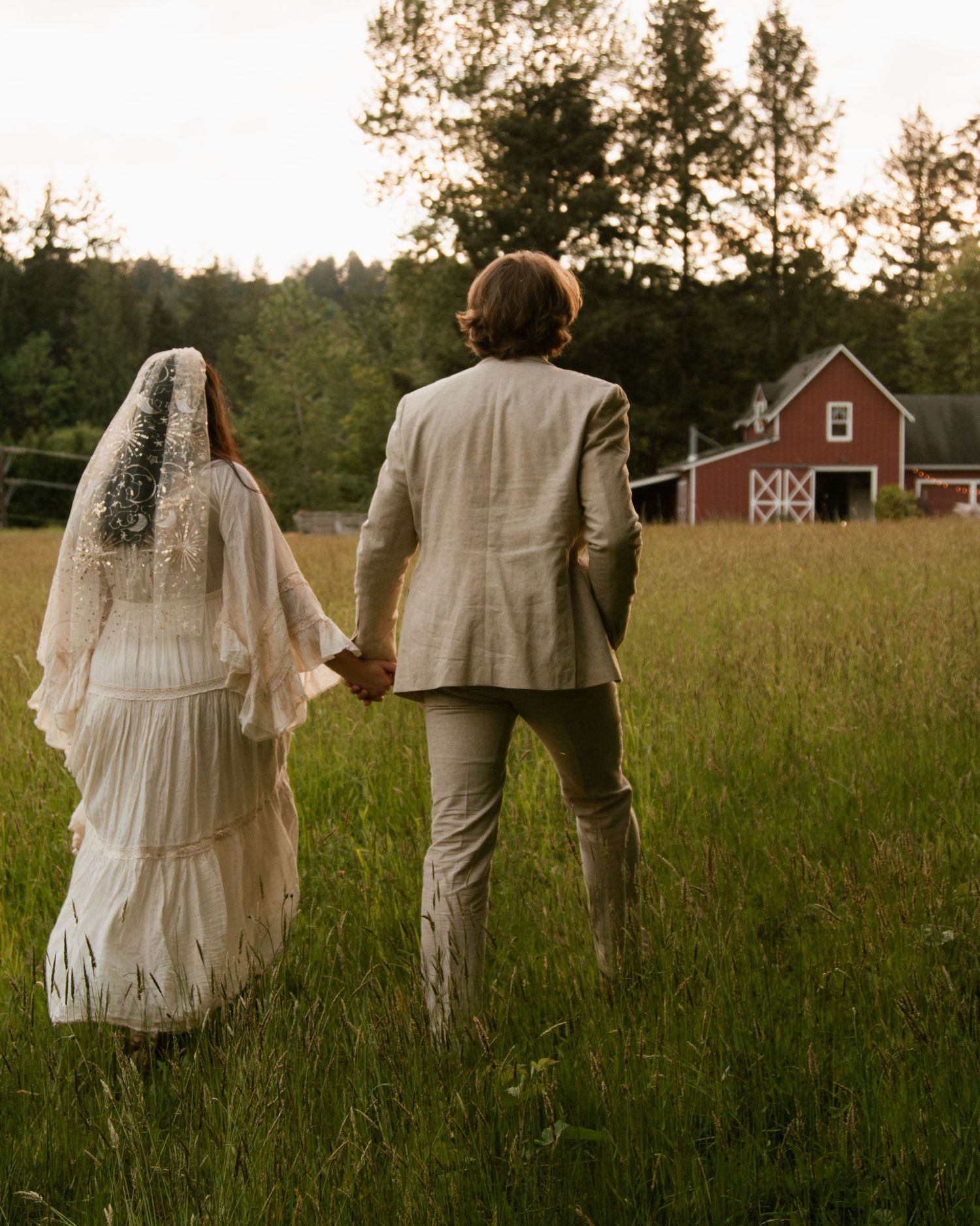 Two figures in light-colored clothing stand in a meadow with a red barn visible in the distance.
