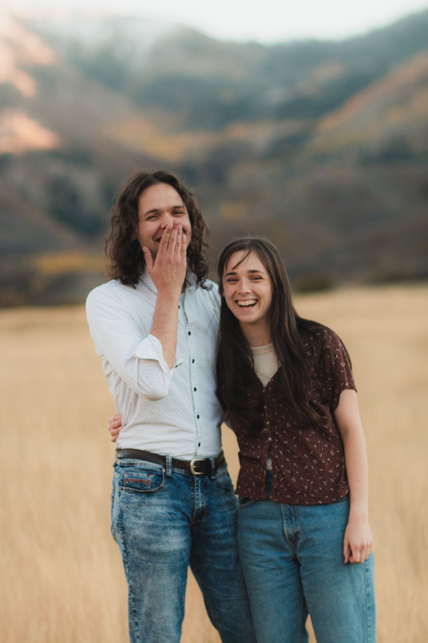 A couple in matching denim jeans and casual tops share candid laughs together in a golden field at sunset.