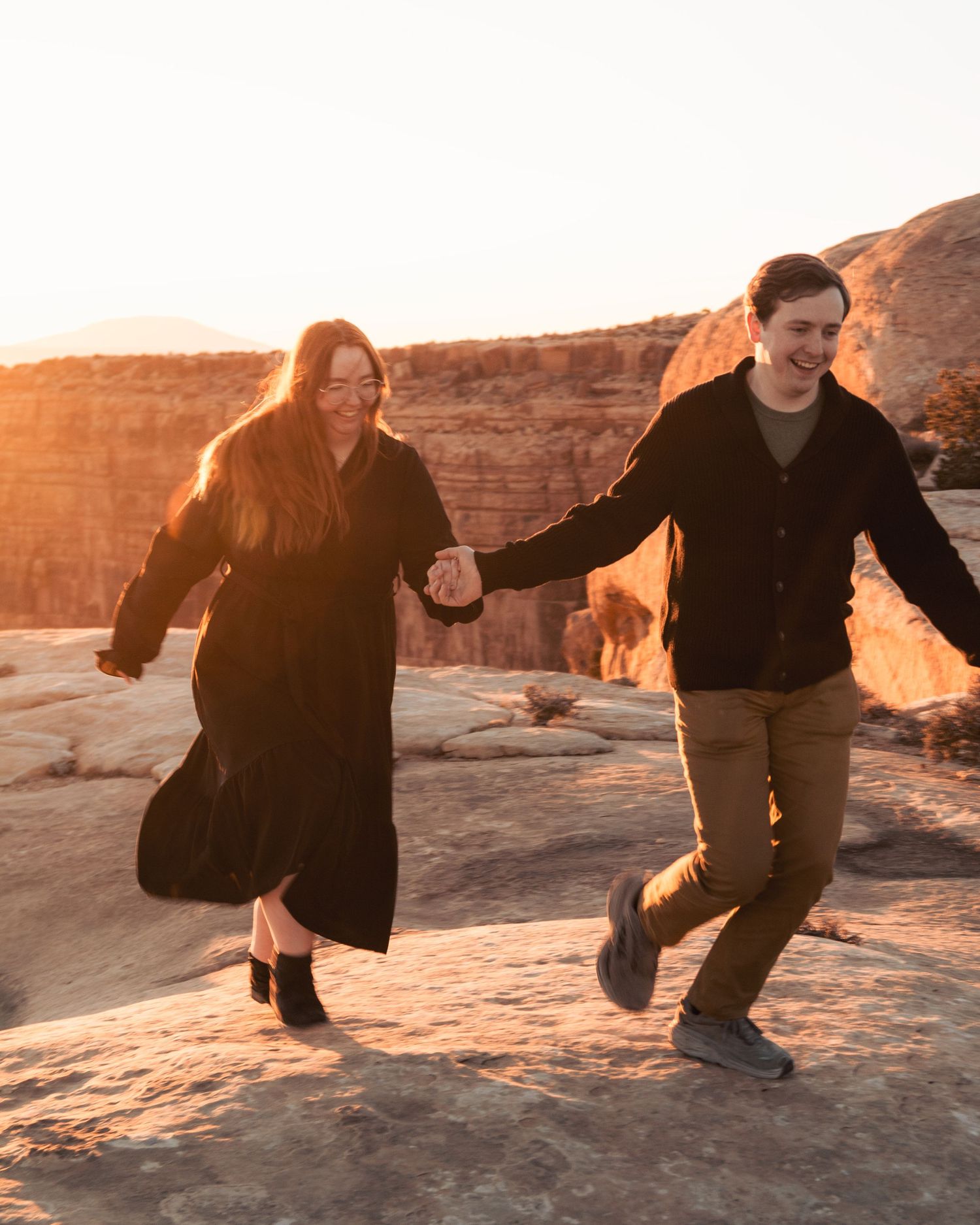 Couple running hand in hand across desert rocks during sunset.