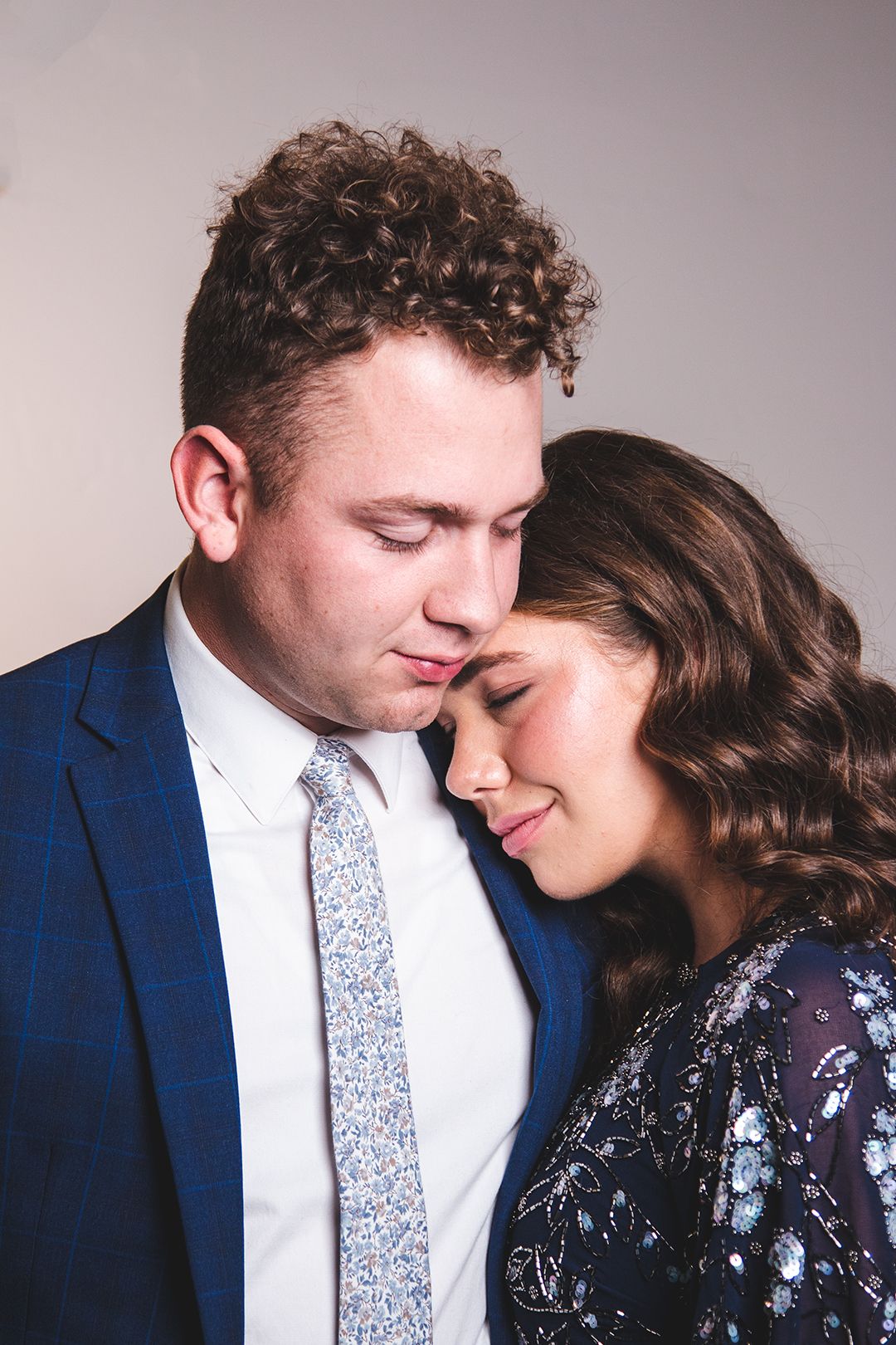 A romantic portrait of a couple in formal attire sharing an intimate moment against a neutral background.