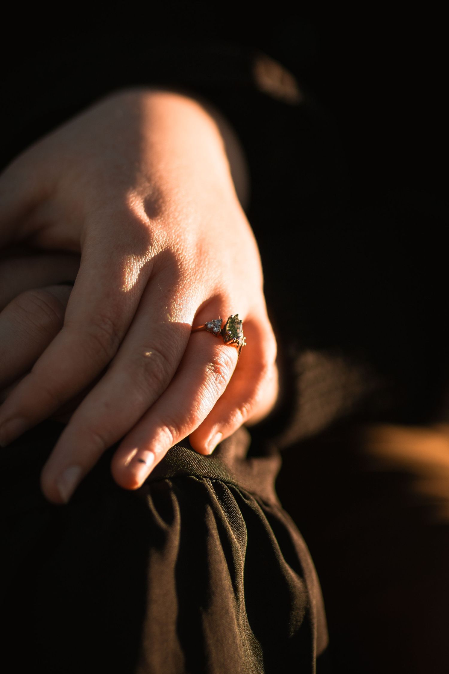 Close-up of hands in sunlight showing an engagement ring against dark fabric.