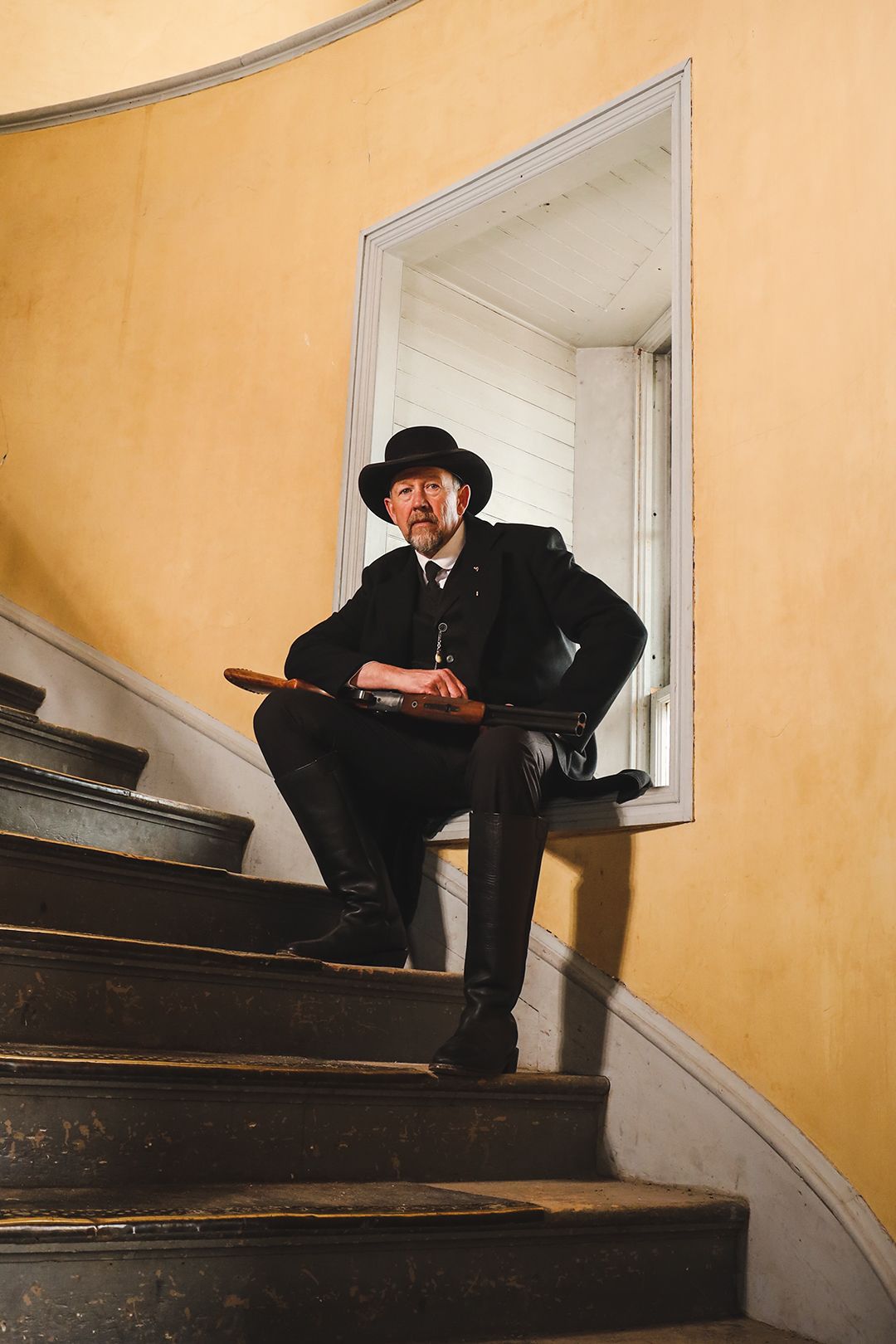 A dark-suited figure with hat poses on curved wooden staircase against yellow wall.