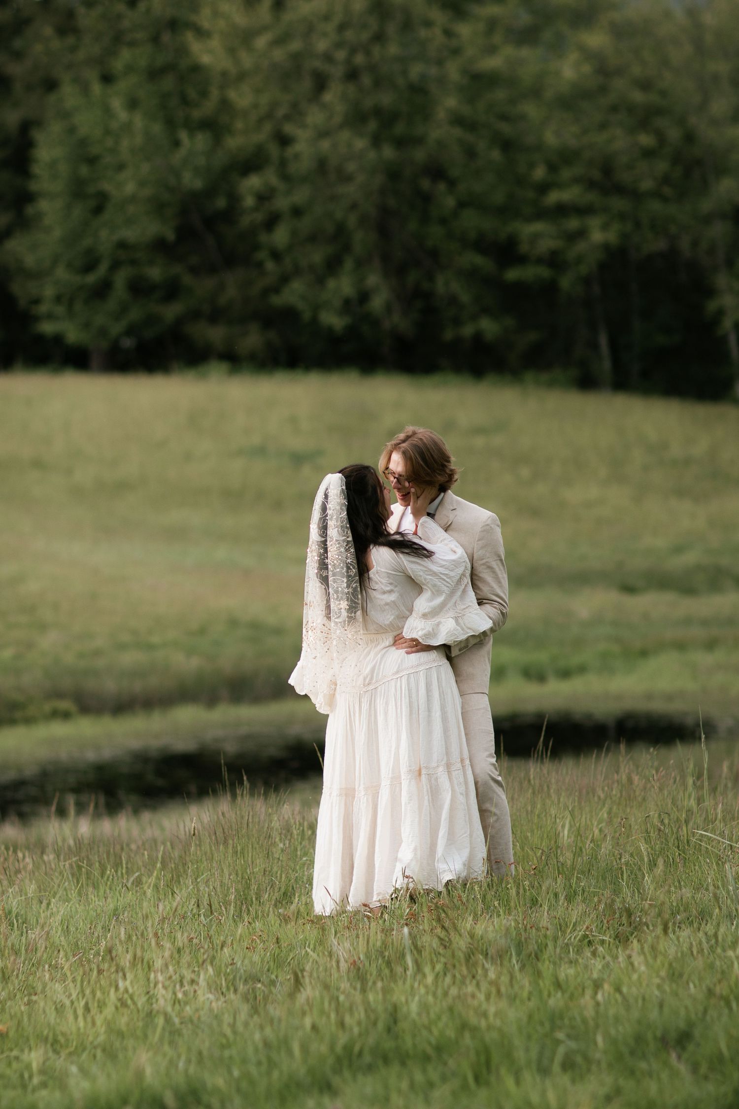 Couple in romantic embrace stands in tall grass under overcast sky.