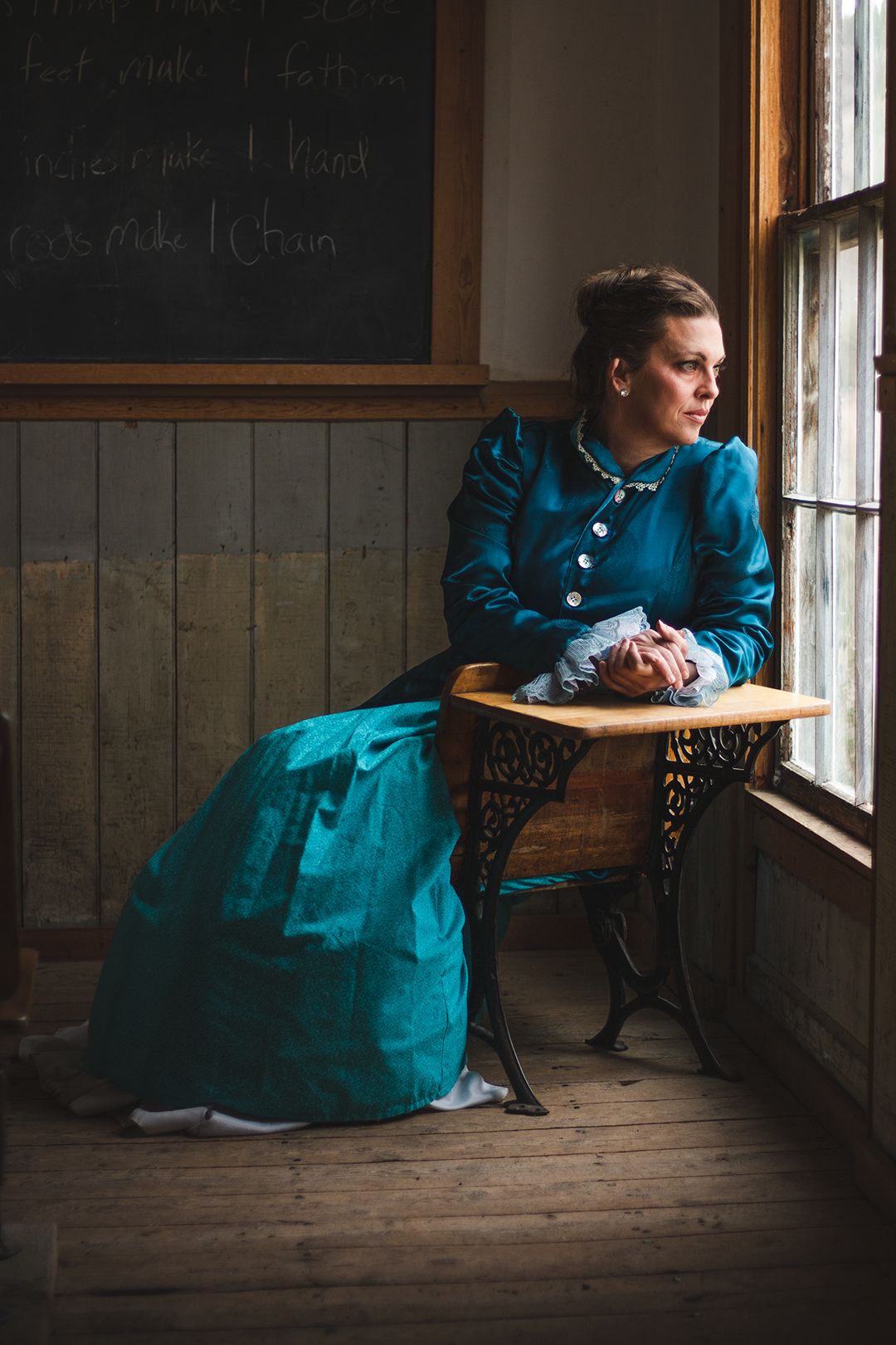 Person in vintage teal dress sitting by window in historic classroom setting.