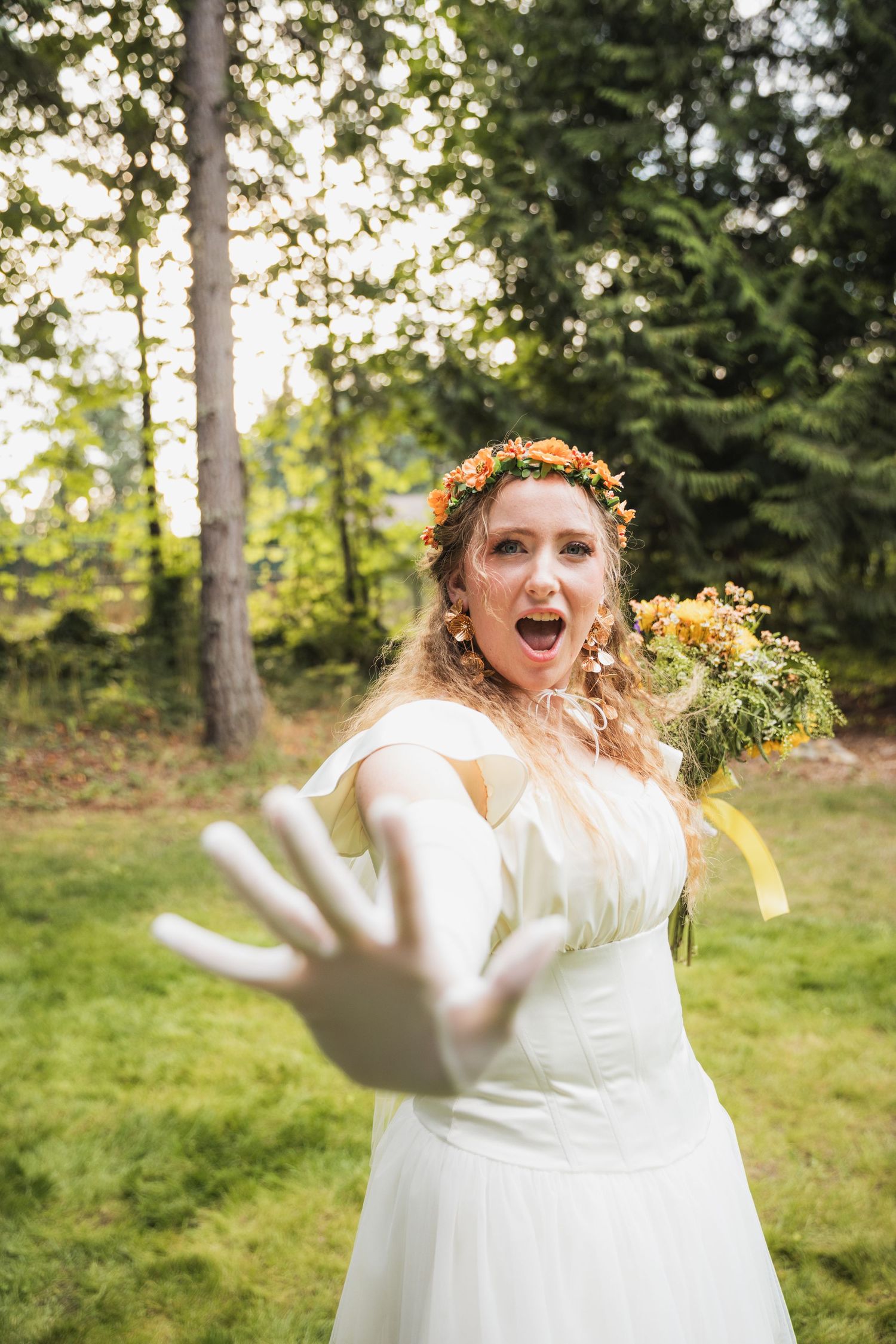 A bride in a white dress and floral crown dances joyfully in a sunlit forest setting.