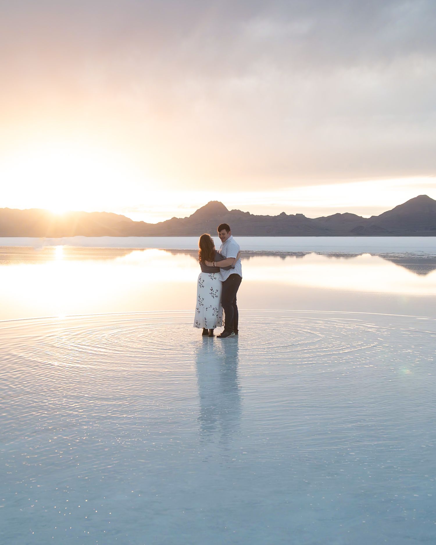 Silhouetted couple embracing on reflective water surface at sunset with mountains.