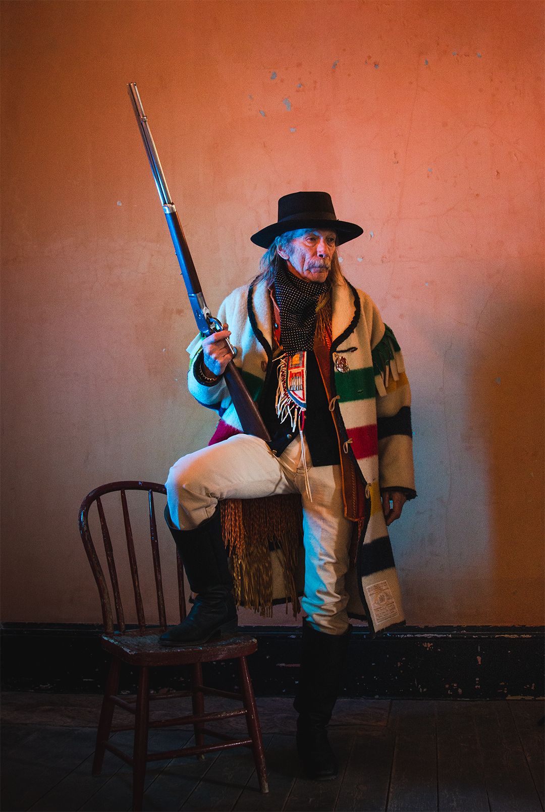 A figure in Western period costume with rifle and hat poses against an orange wall.