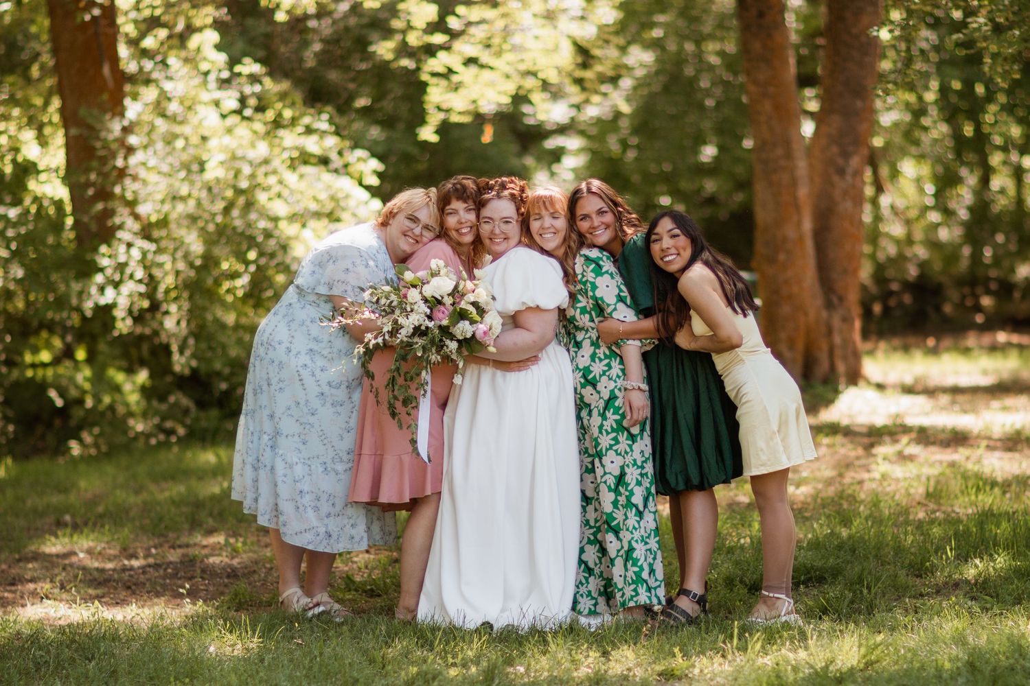 A group of wedding guests in floral dresses pose together under trees in a sunny outdoor setting.