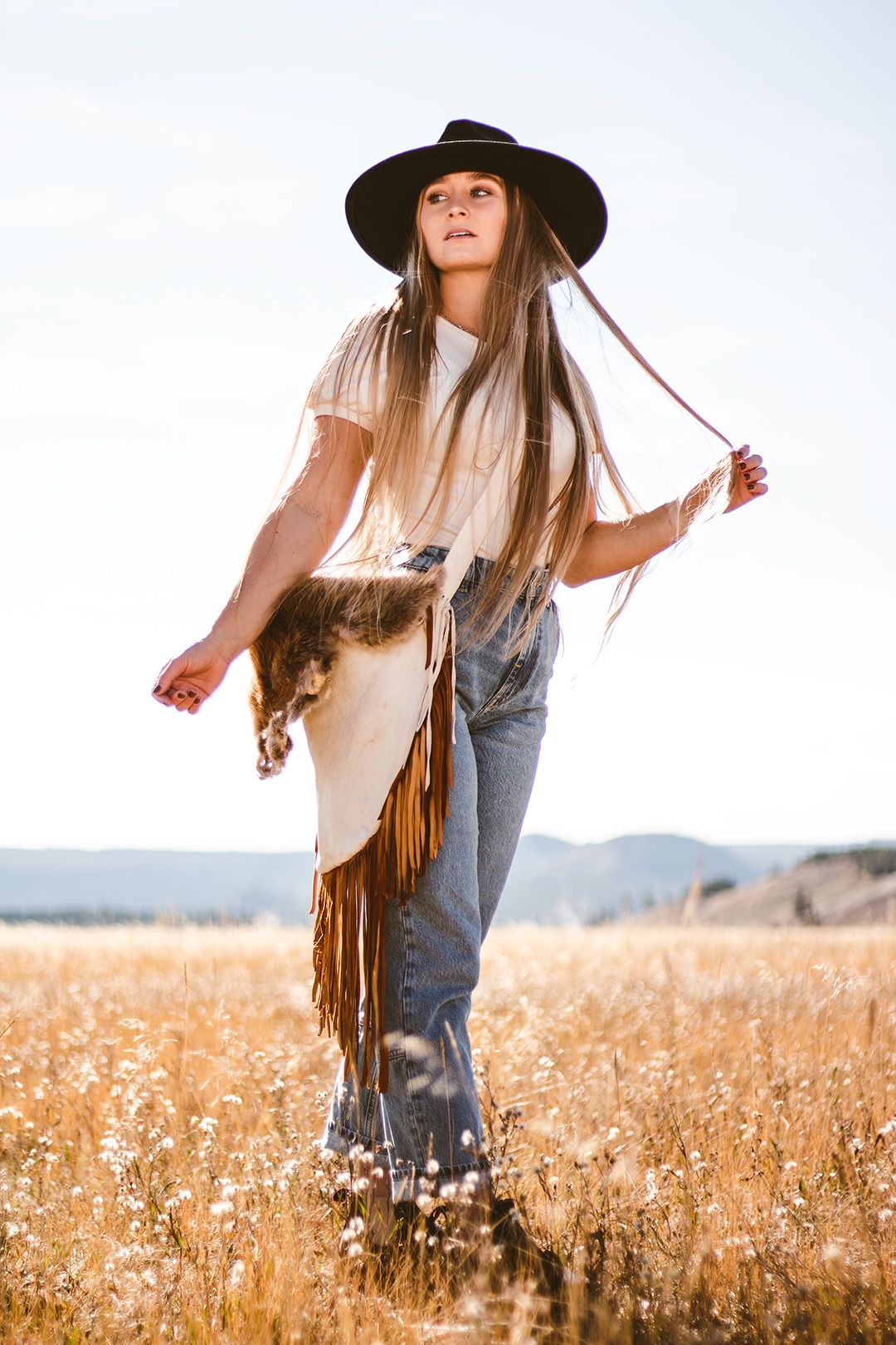 Person in brown hat and fringe vest dancing in golden wheat field at sunset.