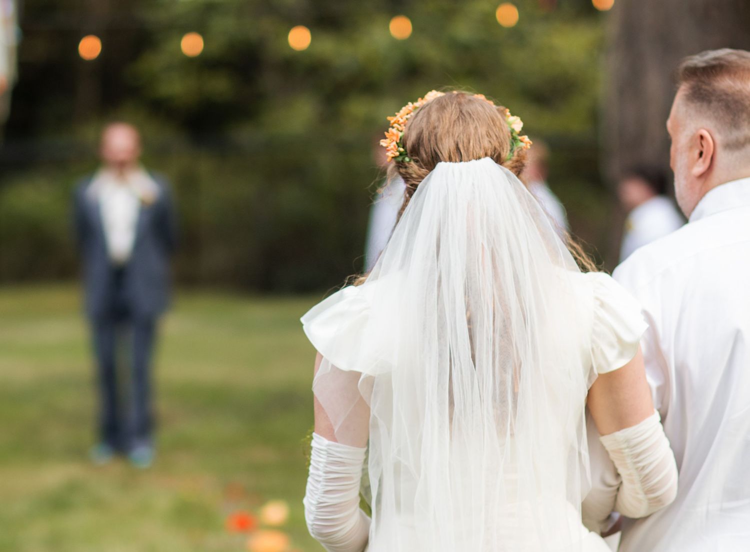 Wedding couple walks hand in hand through a garden setting with glowing string lights overhead at dusk.