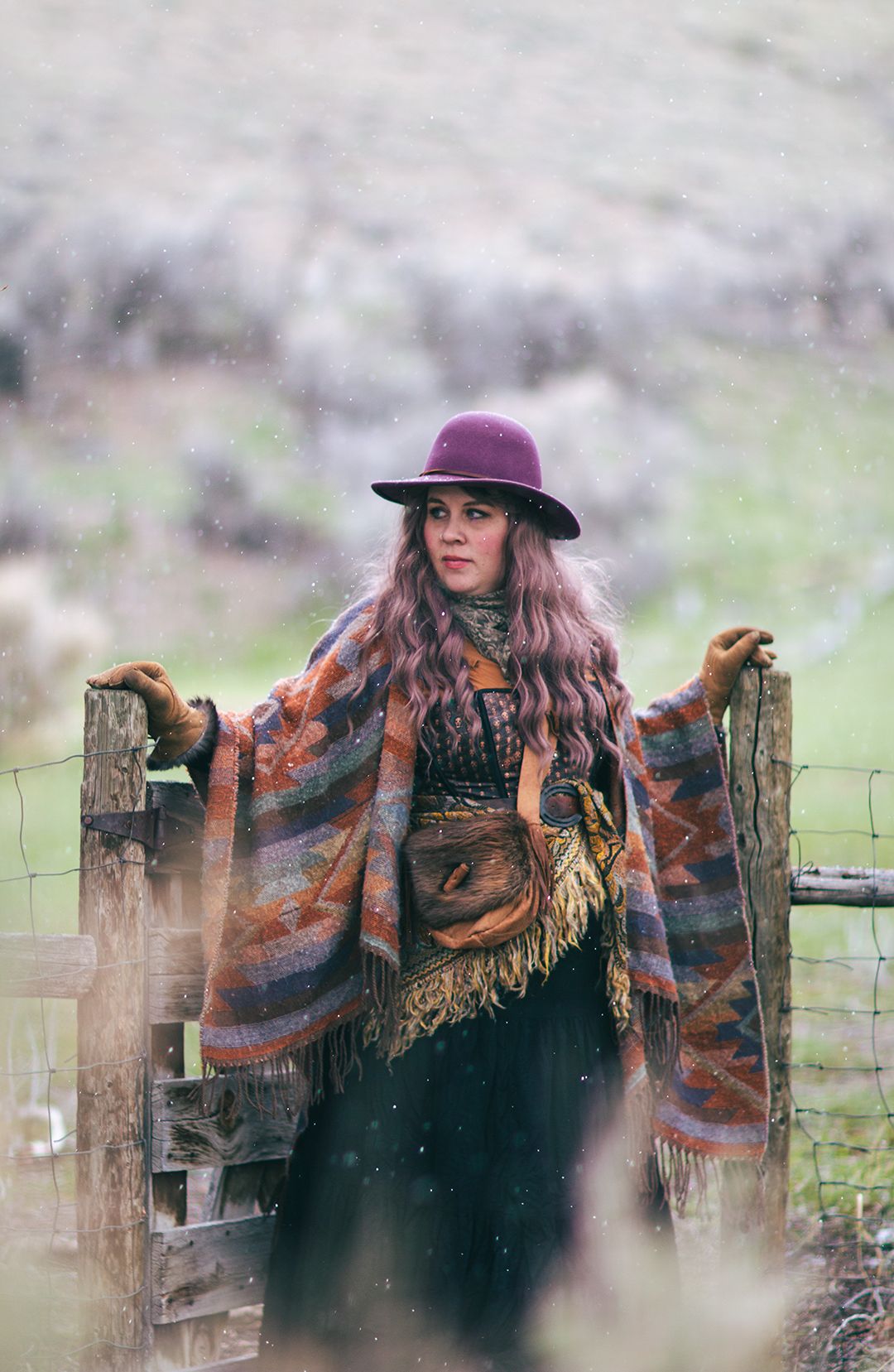 A person in vintage Western attire with purple hat leans against wooden fence in misty weather.