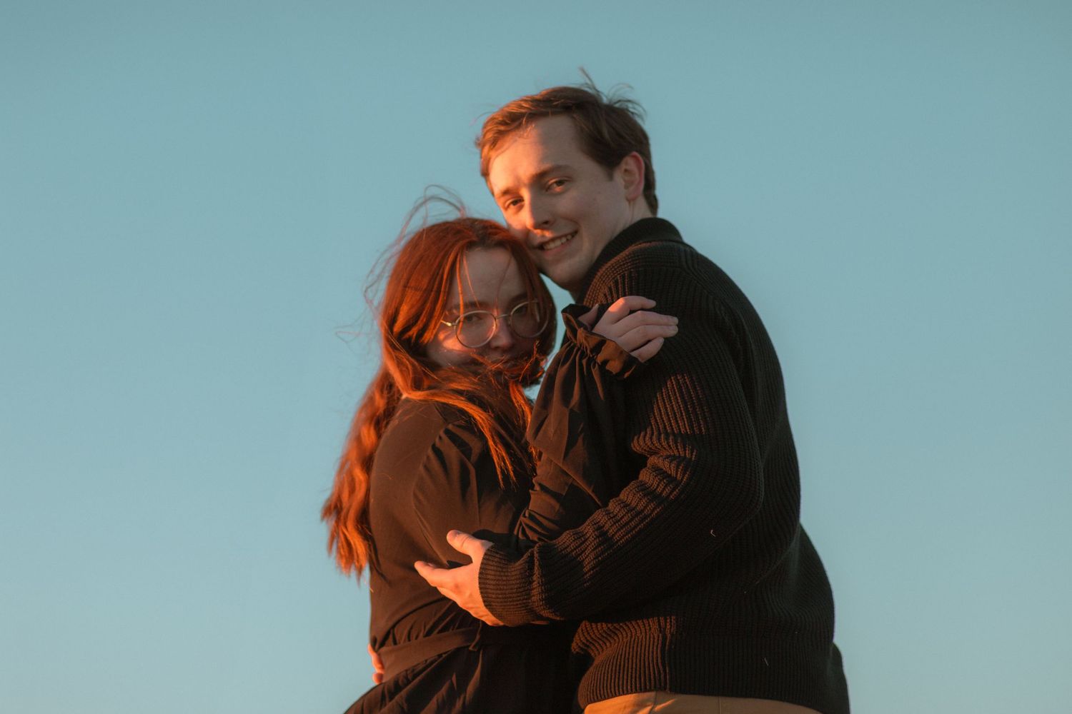 A couple embraces against a blue sky background during golden hour.