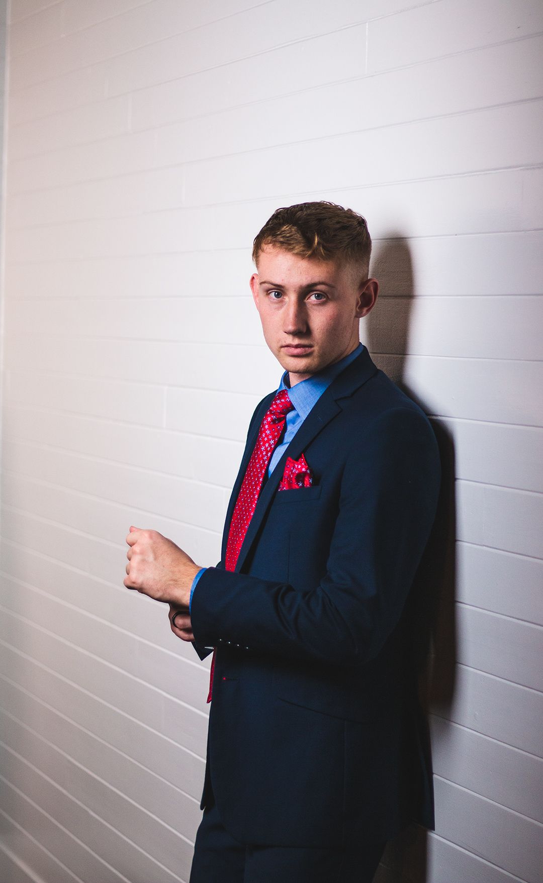 Studio portrait in navy suit and red tie against white wall with dramatic shadow.