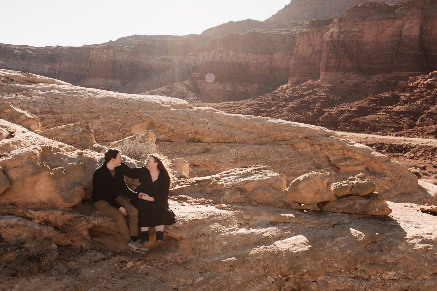Two people sitting together on red rocks in a desert canyon landscape.