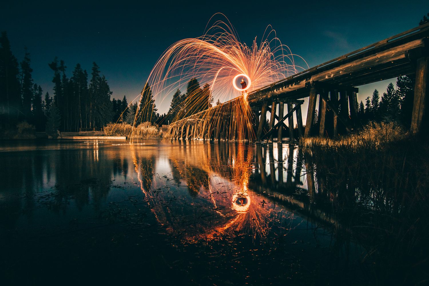 Light painting creates a circular fire pattern reflected in water beneath a wooden bridge at night.