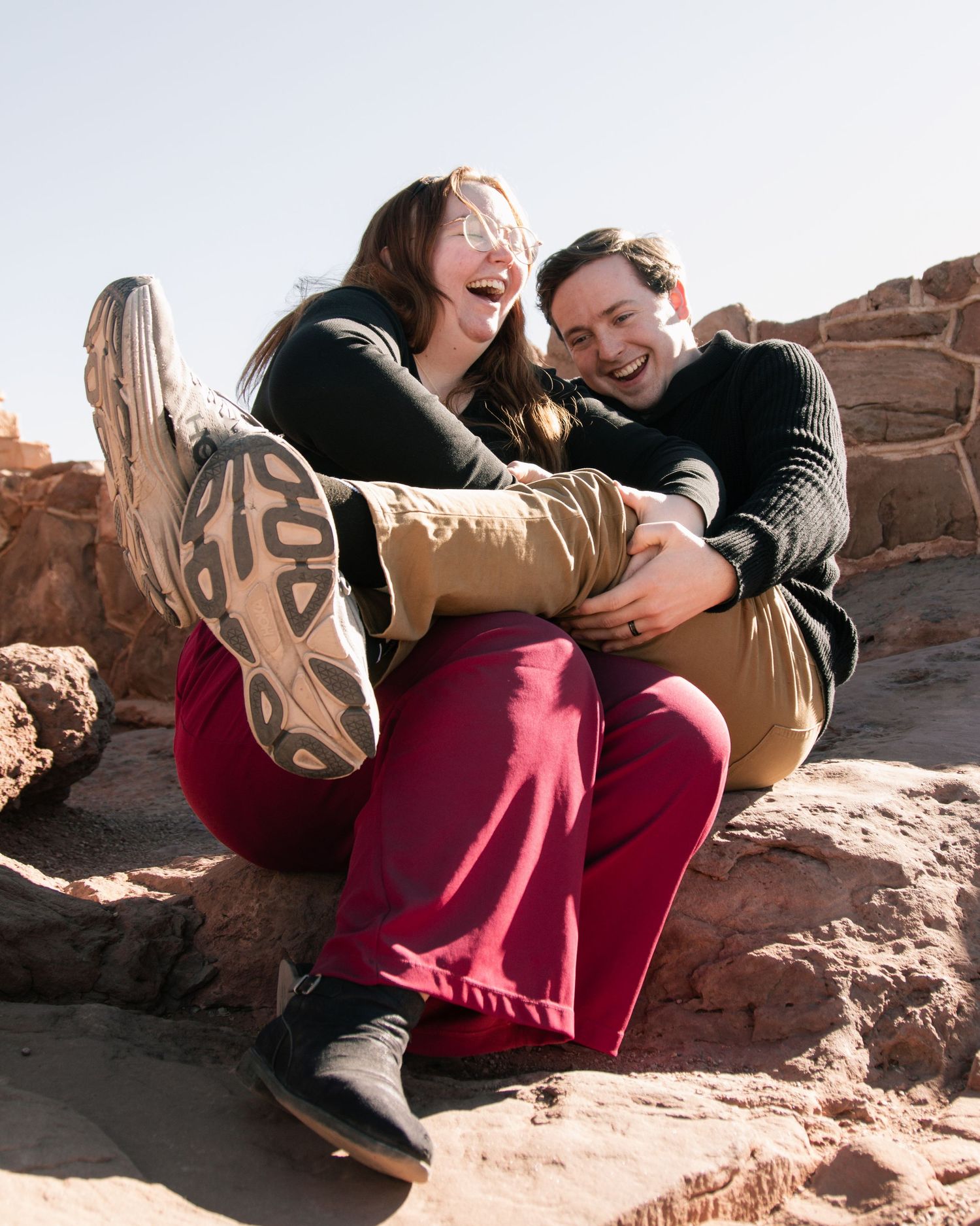 Playful couple shares laughter while resting on red desert rocks.