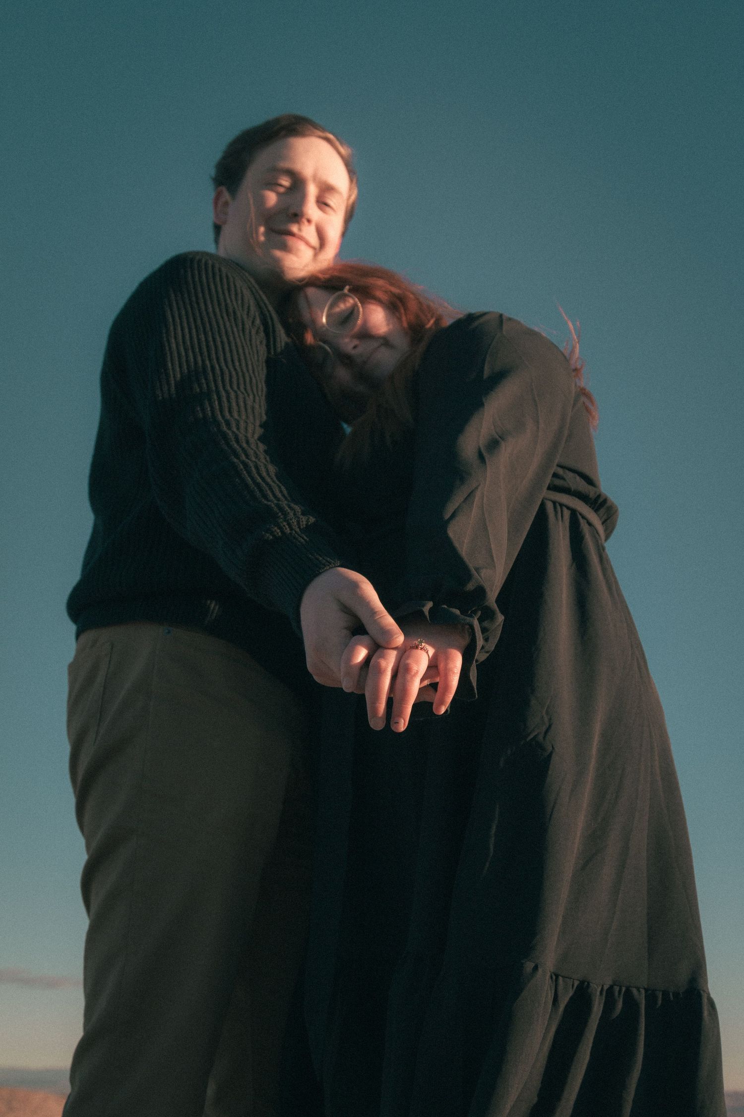 Couple in dark clothing embracing against a teal sunset sky.