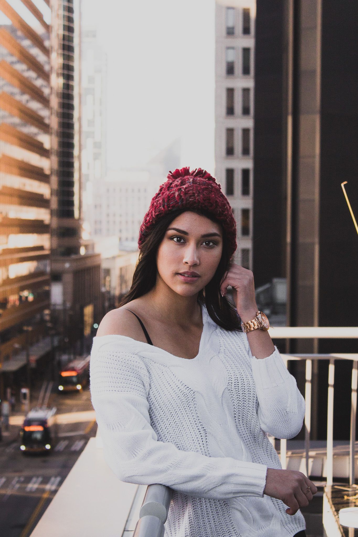 Urban portrait of woman in white sweater and red beanie against city buildings at sunset.