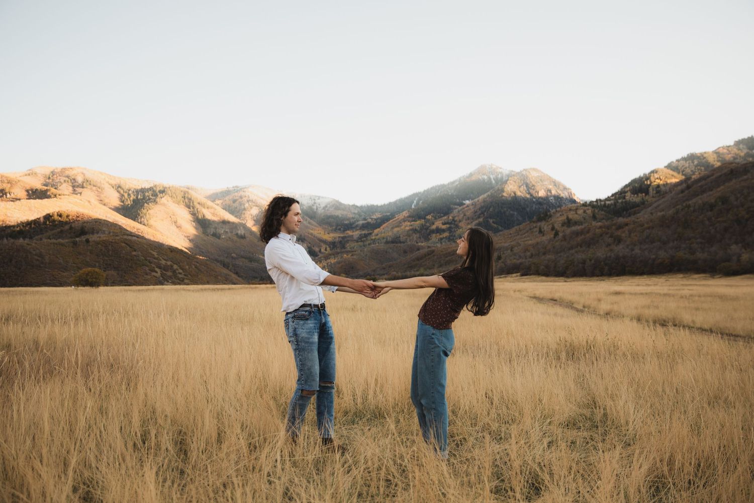 Two people holding hands in a golden grass field with mountains in the background at sunset.