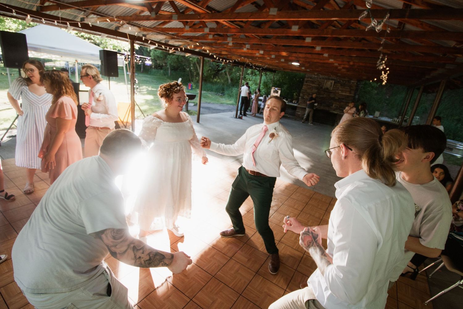 Wedding guests dance and celebrate under a rustic wooden pavilion with natural lighting.