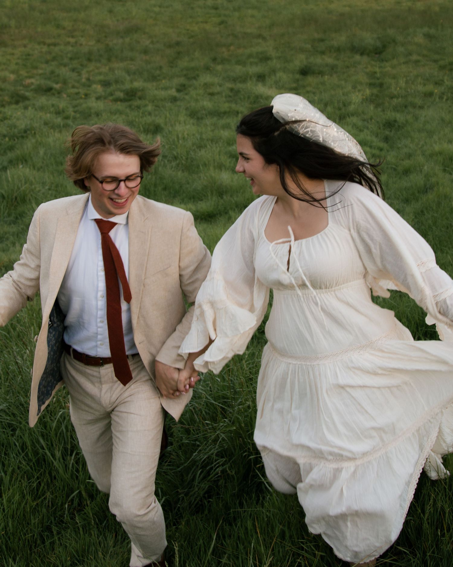 A couple in white flowing attire runs playfully through a grassy field on a sunny day.