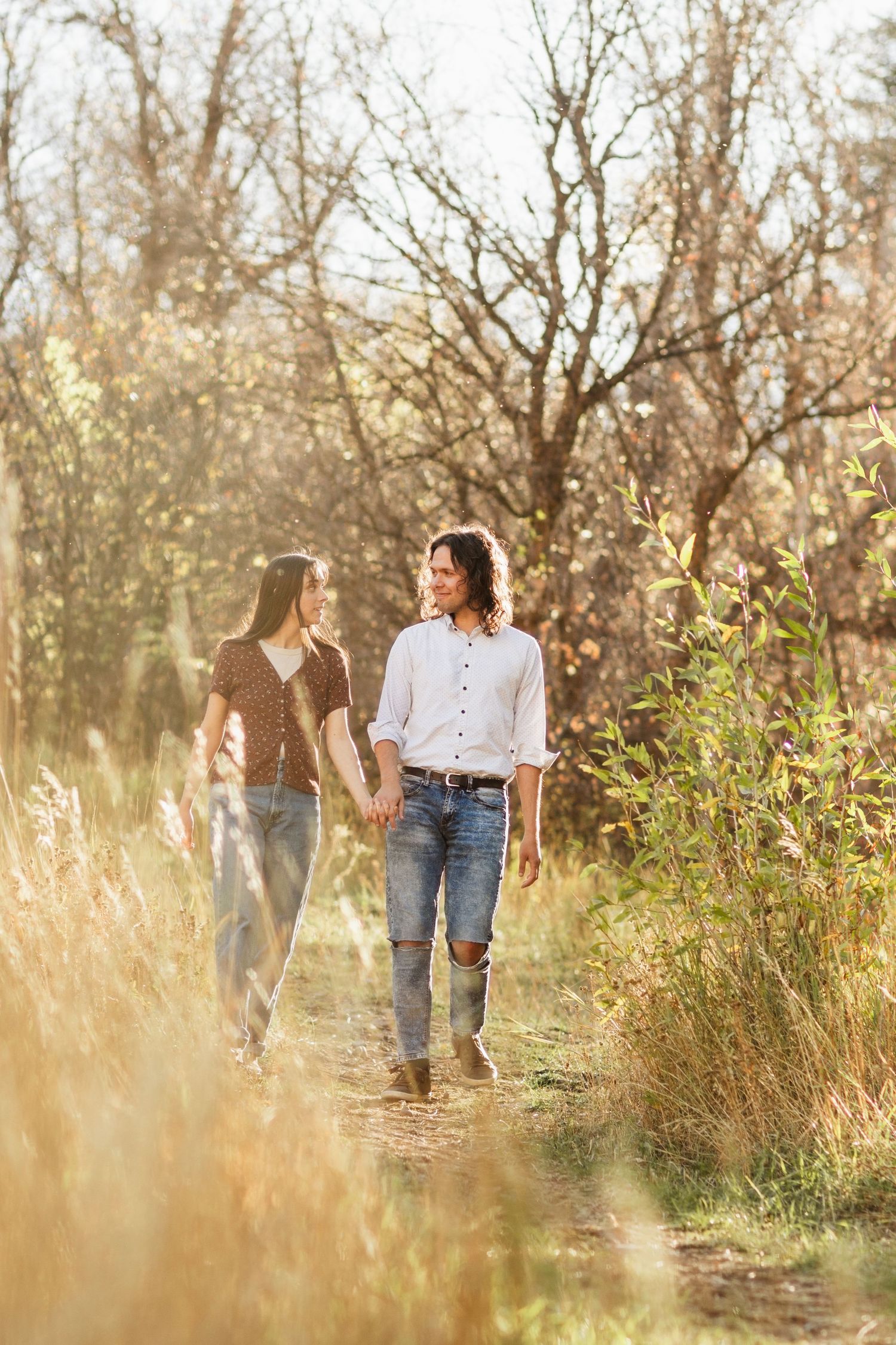 A couple walks hand in hand through tall grass and trees during golden hour sunset lighting.