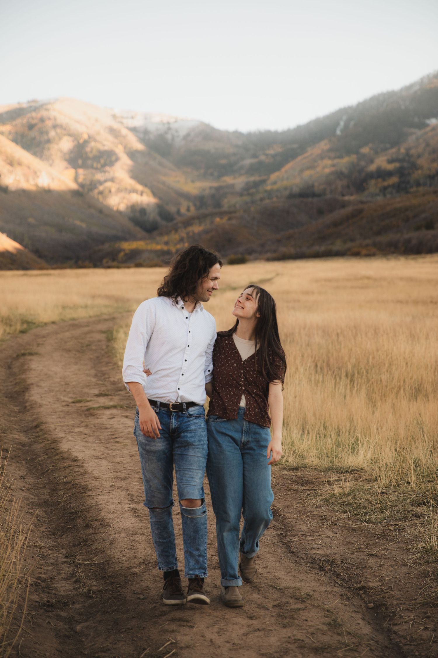 Couple walking together through golden grass fields with mountains in background during sunset.