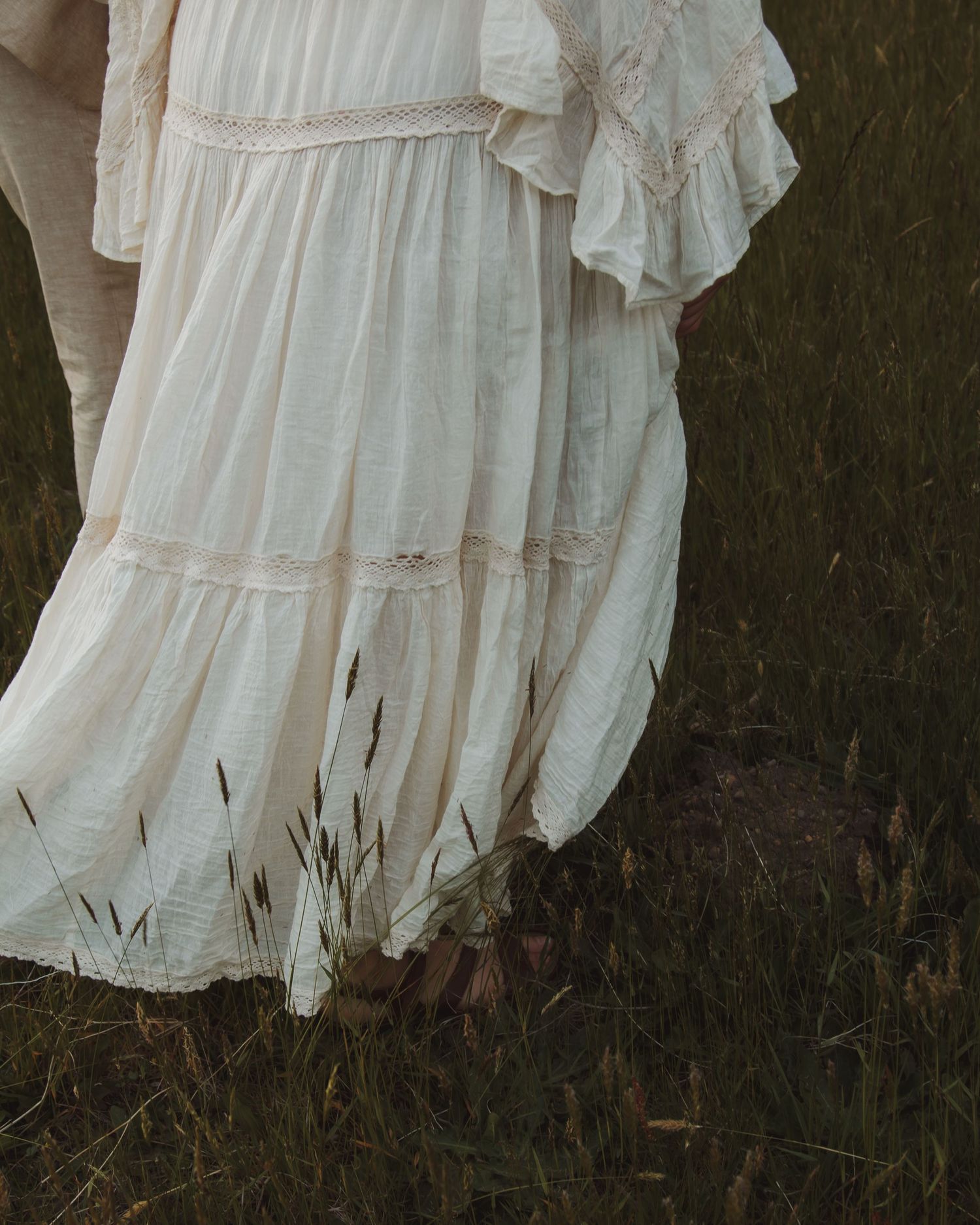 The hem of a flowing white dress rustles against tall grass and cattails.