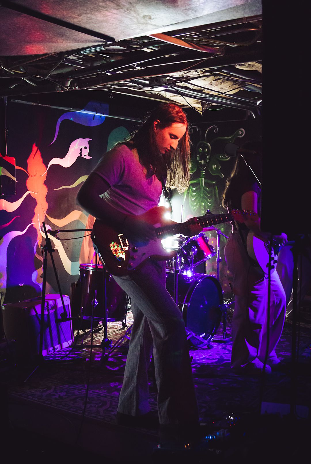 Musician performs with guitar on colorfully lit stage with psychedelic backdrop.