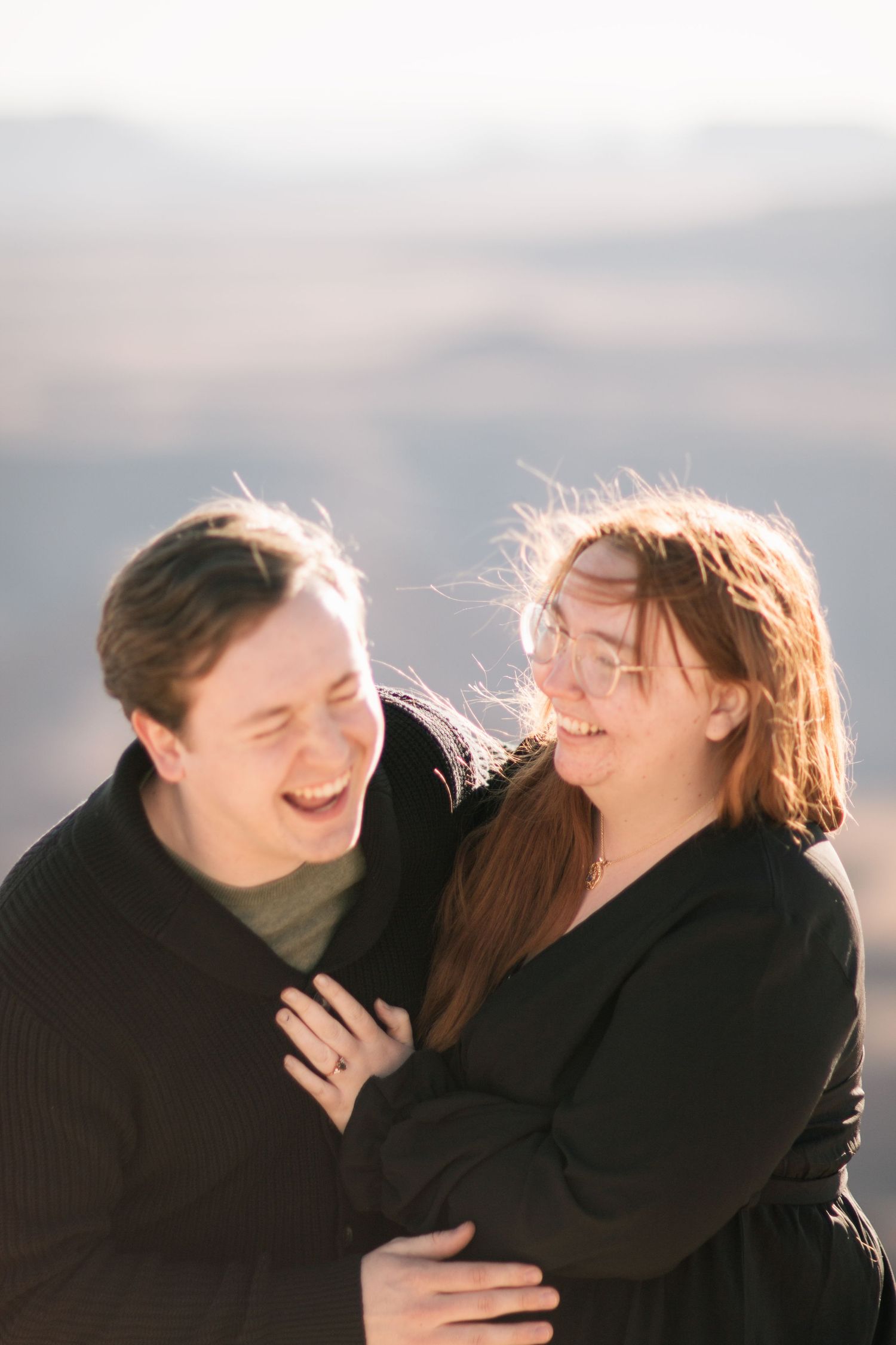 Couple shares joyful laughter together during outdoor photoshoot with natural backlit lighting.