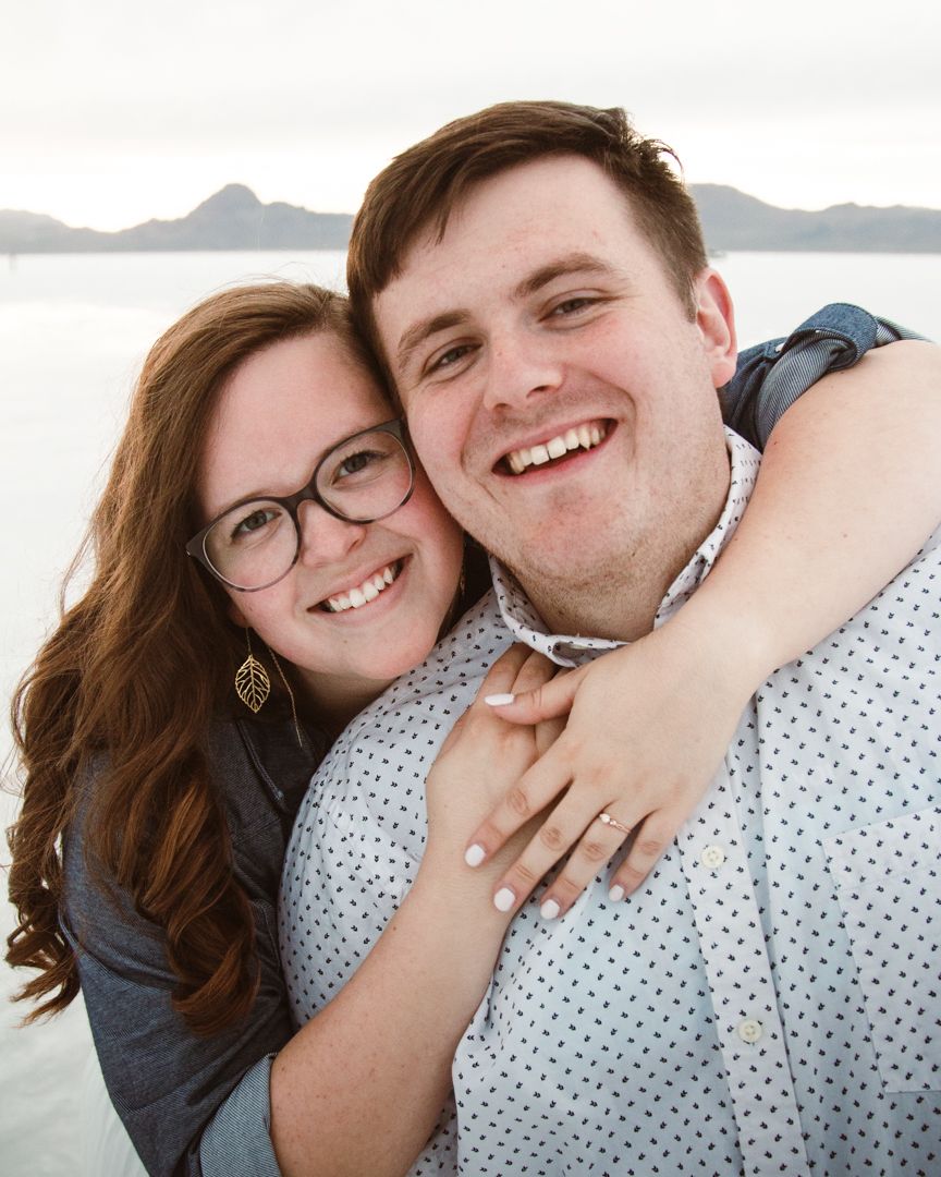 Engaged couple shares a close embrace while smiling at the camera against a mountain backdrop.