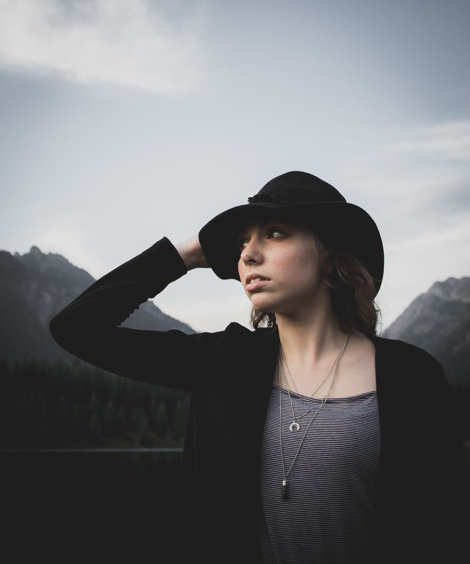Moody fashion portrait in black outfit with wide-brimmed hat against dramatic mountain landscape.