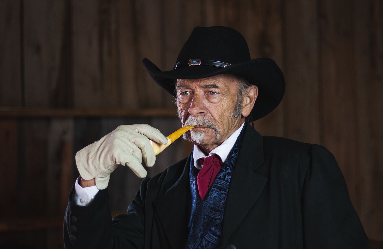 A Western-styled figure in black cowboy hat and formal attire lighting a cigar indoors.