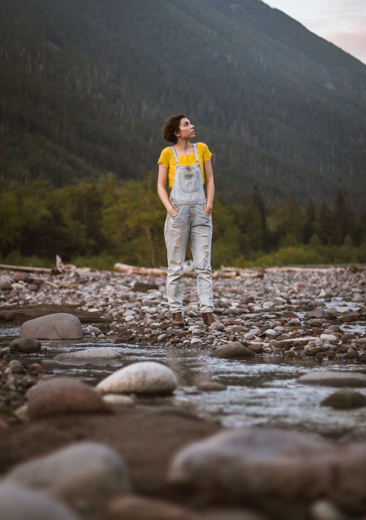 Figure wearing yellow top and overalls standing on rocky riverbed with mountains in background.