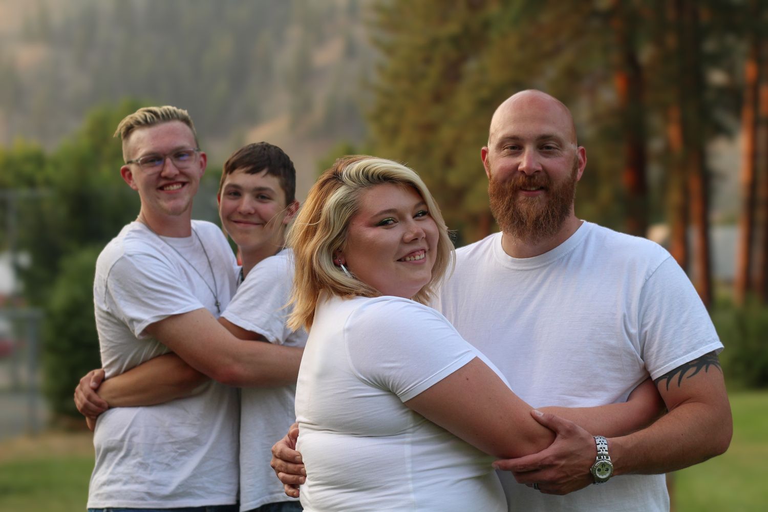 Superior, Mt family portrait pairs hugging 2 adults, 2 teen boys. All are wearing white t-shirts and smiling with pine trees in background.