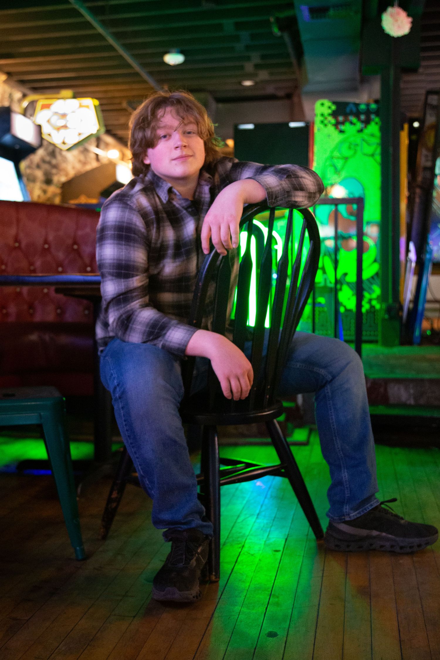 Teen boy at The Guild sitting backward on a chair at an arcade, background has posters of pop culture characters and colorful lights.