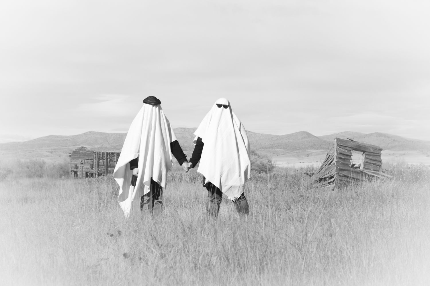 Two people dressed up as ghosts in a field with broken down houses from the turn on the 19th century in the background. Photo is in blk/wht.